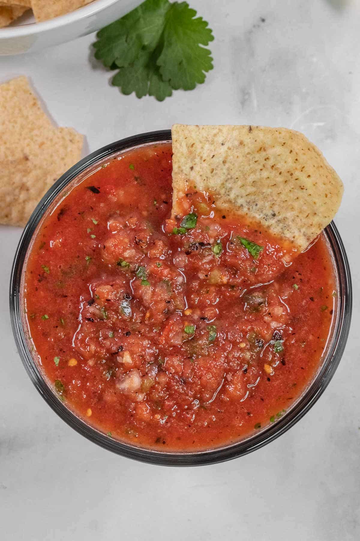 A tortilla chip dipped into a bowl of chunky tomato salsa, with cilantro and other tortilla chips visible in the background.