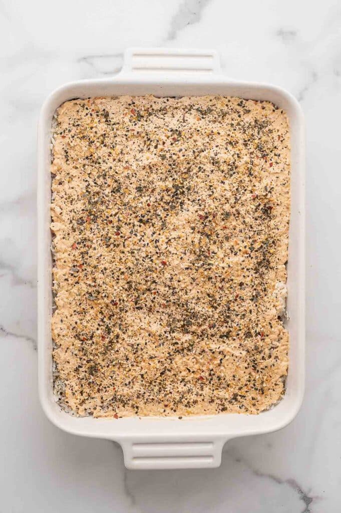 A white baking dish filled with an uncooked spicy sushi bake mixture topped with furikake seasoning.