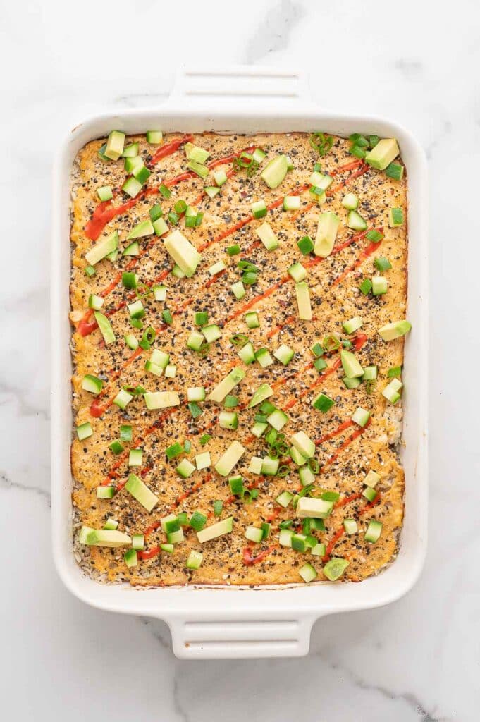 A rectangular casserole dish filled with cooked sushi bake, drizzled with sriracha and topped with cucumber.