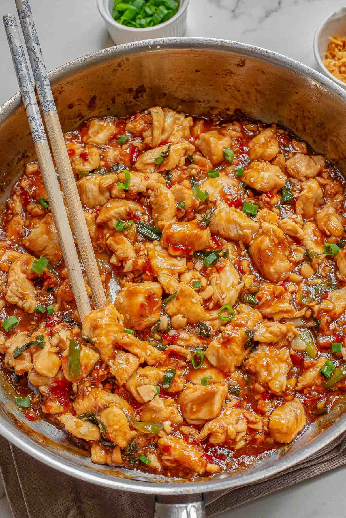 A skillet filled with cooked diced chicken in a sweet chili sauce, garnished with green onions with chopsticks on the side.