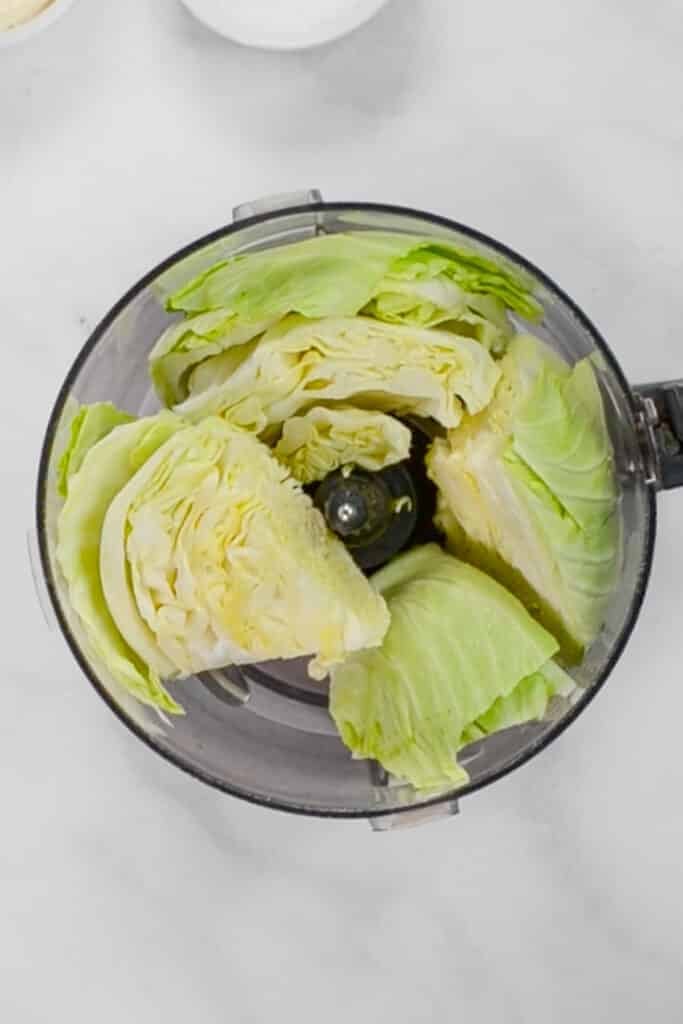 Overhead view of cabbage wedges placed inside a food processor bowl on a white surface.