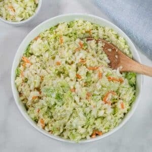 A bowl of creamy coleslaw with shredded cabbage and carrots, mixed with dressing, and a wooden spoon resting inside.