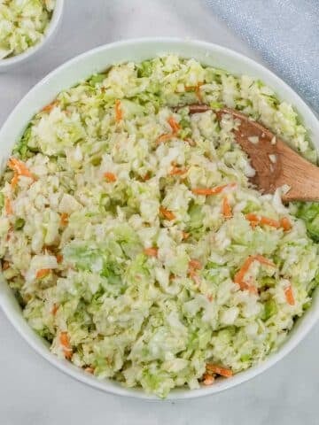 A bowl of creamy coleslaw with shredded cabbage and carrots, mixed with dressing, and a wooden spoon resting inside.