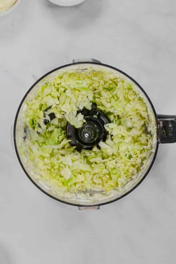 Chopped cabbage pieces inside a food processor viewed from above on a light-colored surface.