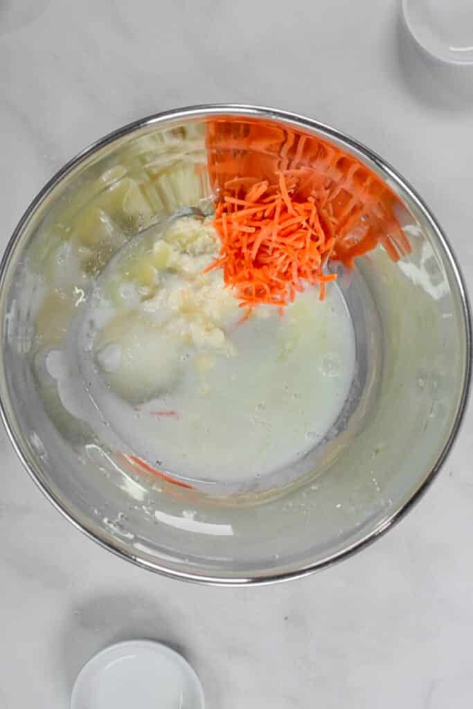 A stainless steel mixing bowl contains shredded carrots, milk, and a white mixture for coleslaw dressing on a light surface.