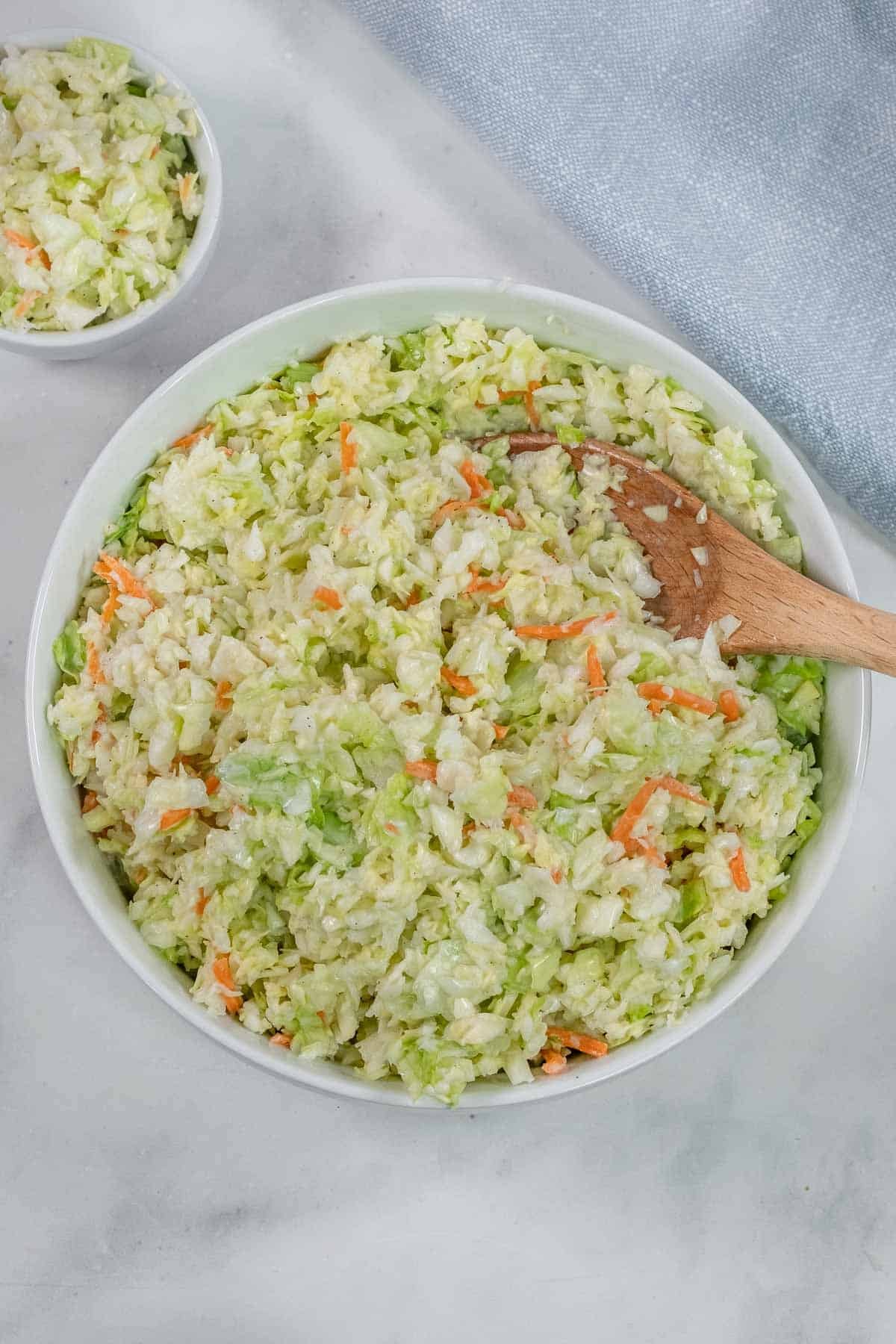 A bowl of an easy coleslaw recipe with finely shredded cabbage and carrots, mixed with a creamy dressing, sits on a light surface with a wooden spoon.