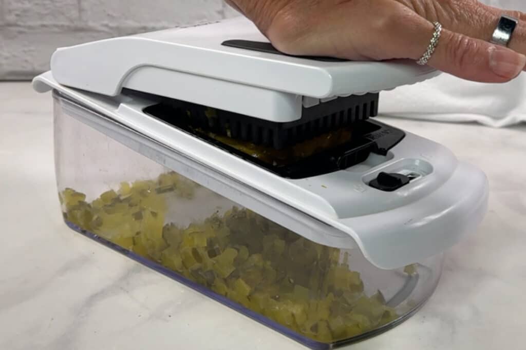 A hand presses down on a vegetable chopper, dicing ingredients for dill pickle dip into small cubes that collect in the attached container.