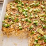 An easy sushi bake casserole recipe topped with diced avocado, sliced green onions, and seasoning, with a corner piece removed to reveal layers of rice and filling.