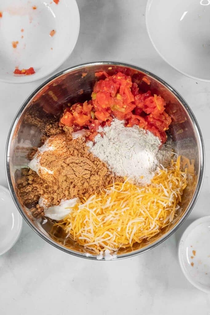 A metal mixing bowl containing shredded cheese, diced tomatoes, ranch mix, taco seasoning, and sour cream ready to be mixed together.