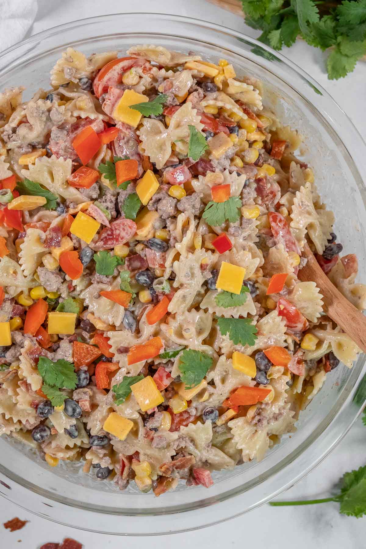 A clear glass bowl filled with cowboy pasta salad on a white surface with a wooden spoon inside.
