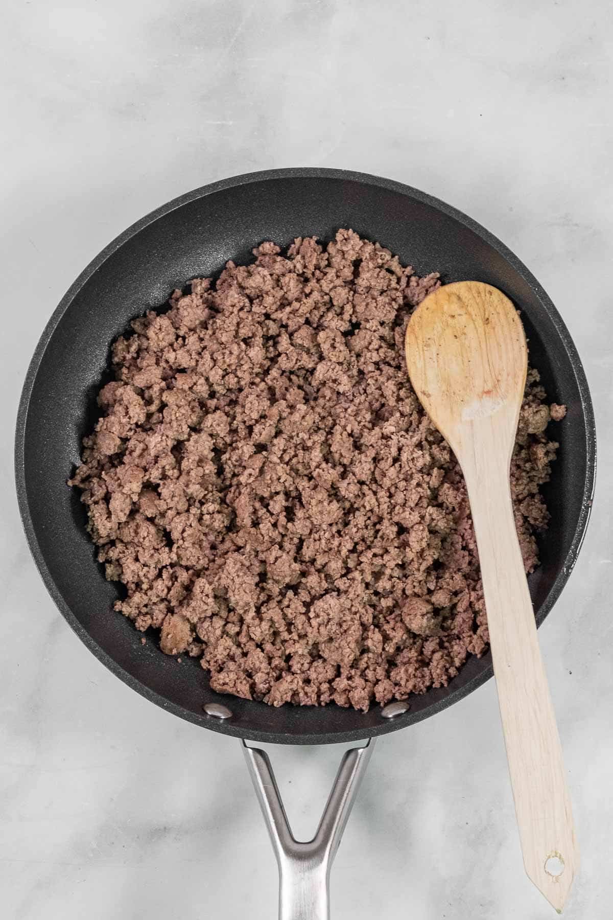 A frying pan with cooked ground beef and a wooden spoon.