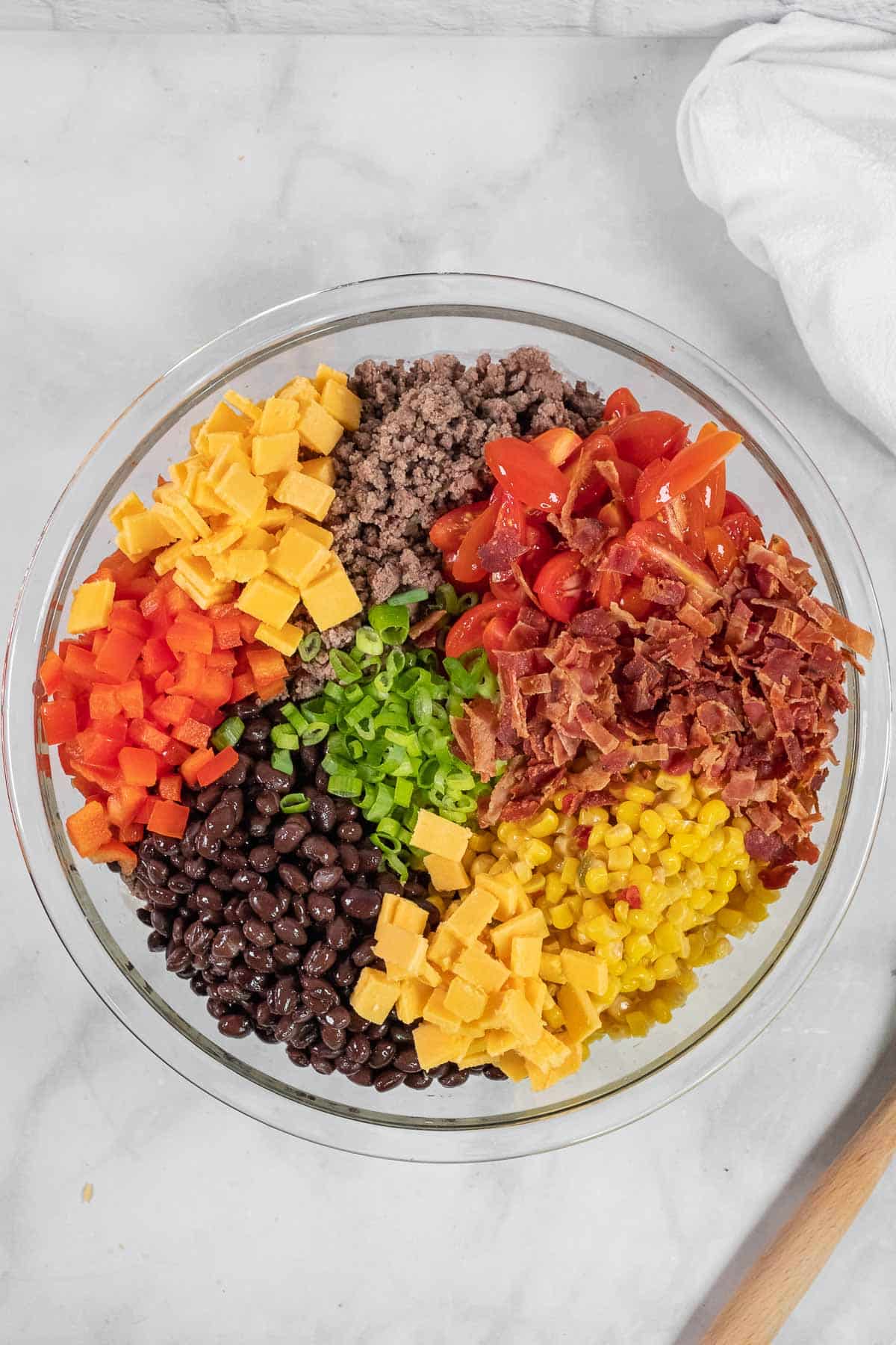 A glass bowl filled with ground beef, cherry tomatoes, bacon, cheddar cheese cubes, corn, black beans, chopped green onions, and red bell pepper, arranged in sections.