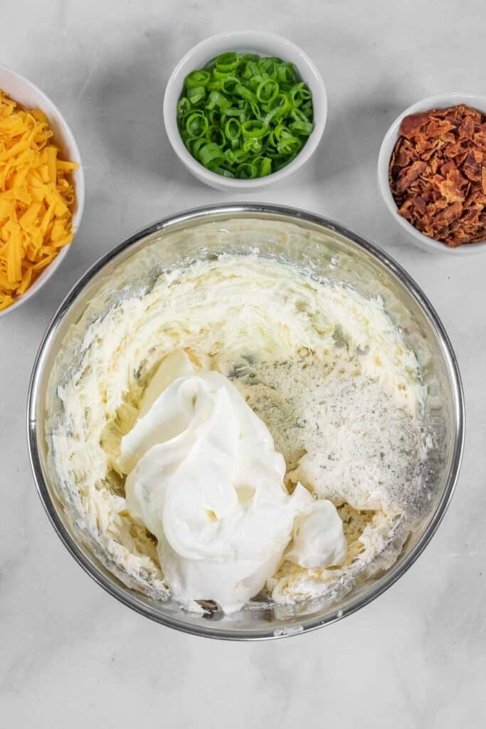 A glass bowl with cream cheese, sour cream, and seasoning being mixed, surrounded by small bowls of shredded cheese, chopped green onions, and bacon bits.