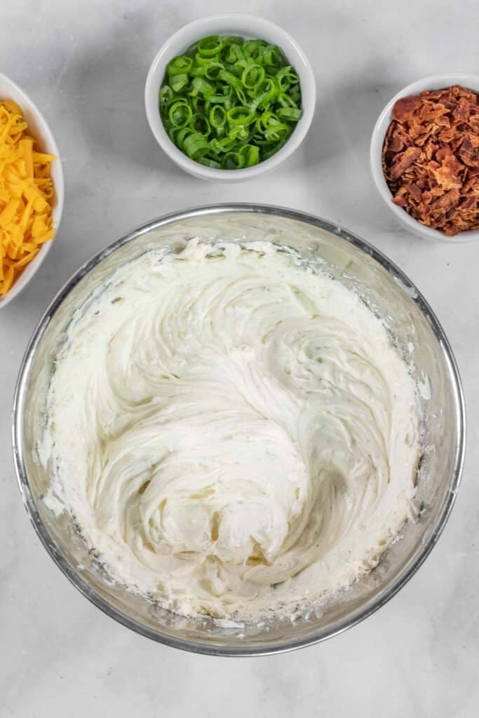 A glass bowl with cream cheese, sour cream, and seasoning being mixed, surrounded by small bowls of shredded cheese, chopped green onions, and bacon bits.