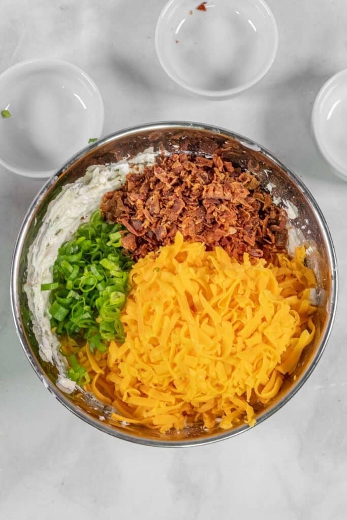 A metal bowl containing chopped green onions, shredded cheddar cheese, crumbled bacon over the cream cheese base.