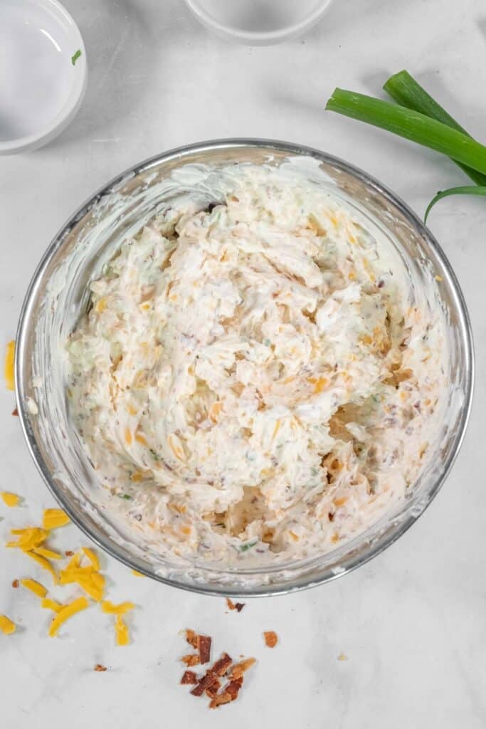 A glass bowl filled with a creamy mixture of shredded cheese, bacon, and herbs sits on a white surface.