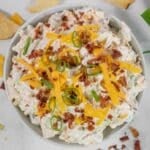 A bowl of creamy crack dip with shredded cheddar cheese, bacon bits, and sliced green onions on top.