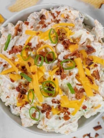 A bowl of creamy crack dip with shredded cheddar cheese, bacon bits, and sliced green onions on top.