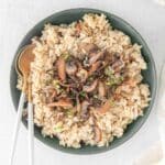 A bowl filled with mushroom risotto garnished with chopped herbs, with golden spoons on the side.