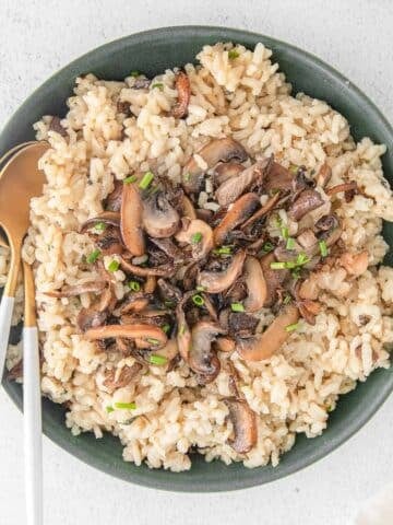 A bowl filled with mushroom risotto garnished with chopped herbs, with golden spoons on the side.