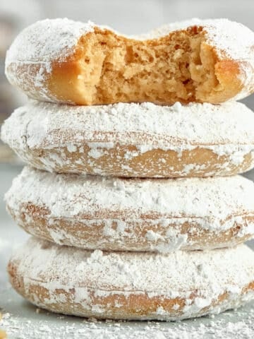 A close up of a stack of four powdered sugar donuts with a bite taken out of the top donut.