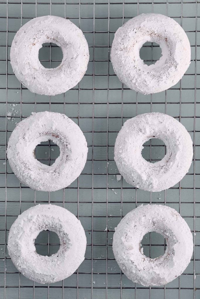 Six powdered sugar donuts are arranged in two vertical columns on a metal cooling rack.