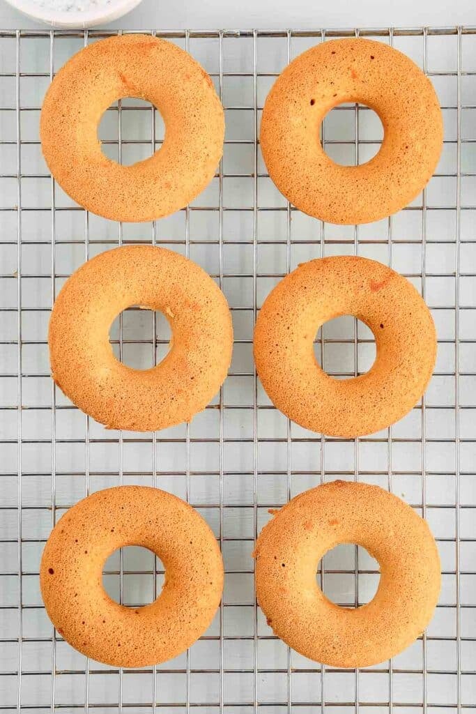 Six plain, baked donuts cooling on a metal wire rack, arranged in two columns of three on a light-colored surface.