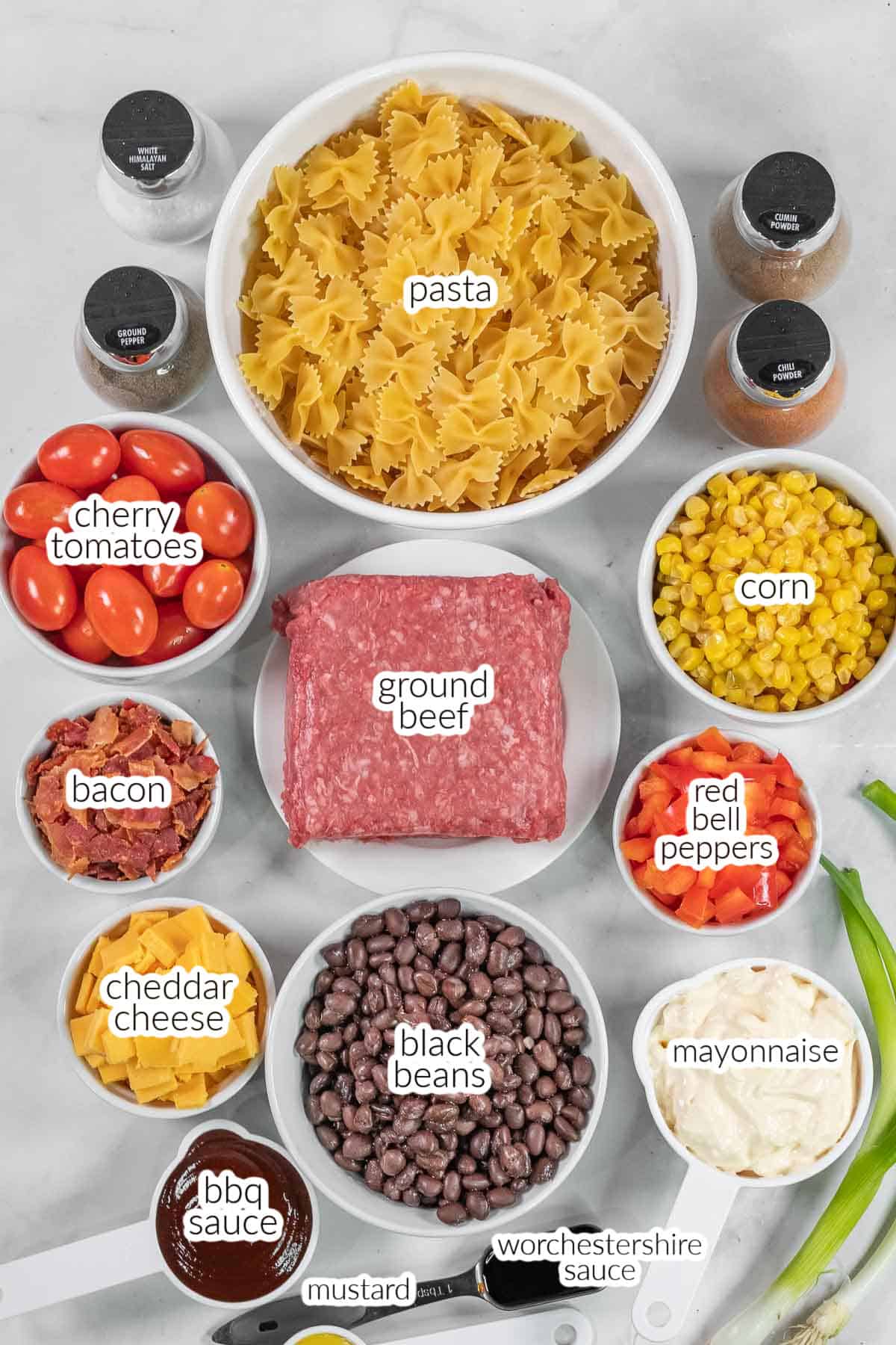A variety of labeled ingredients for Cowboy Pasta Salad on a counter, including pasta, ground beef, black beans, corn, cherry tomatoes, bacon, cheddar cheese, bell peppers, mayonnaise, Worcestershire sauce, mustard and bbq sauce.