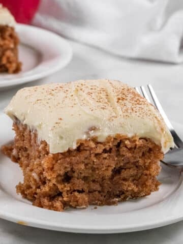 A slice of frosted applesauce cake with a dusting of cinnamon is served on a white plate with a fork.