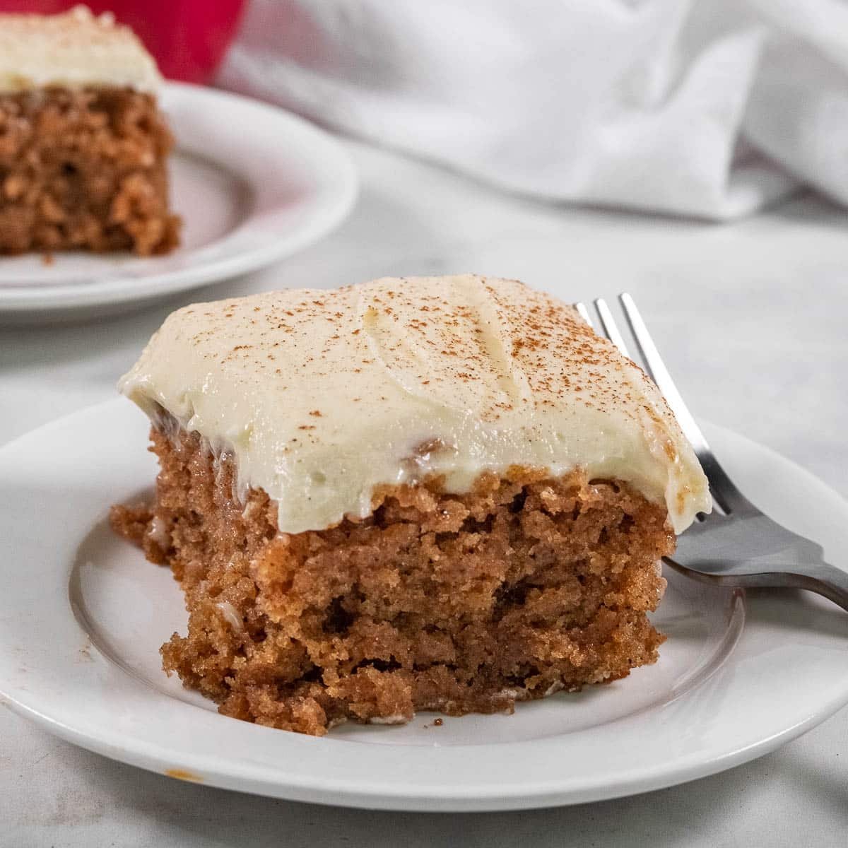 A slice of frosted applesauce cake with a dusting of cinnamon is served on a white plate with a fork.
