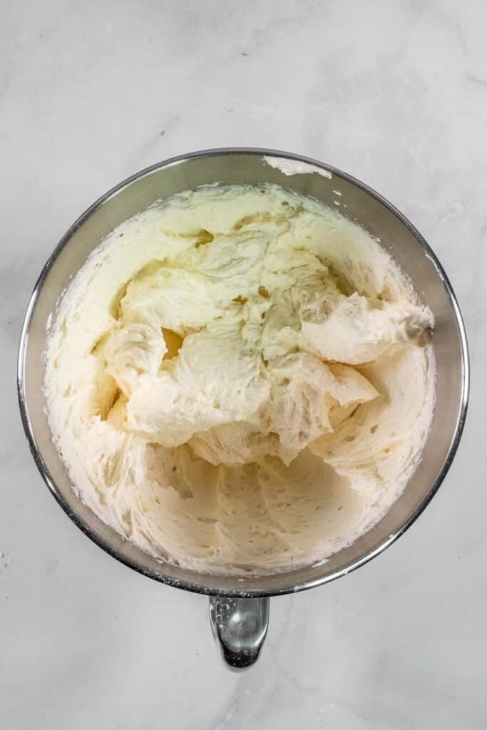A metal mixing bowl filled with whipped, fluffy white buttercream frosting sits on a light marble surface.