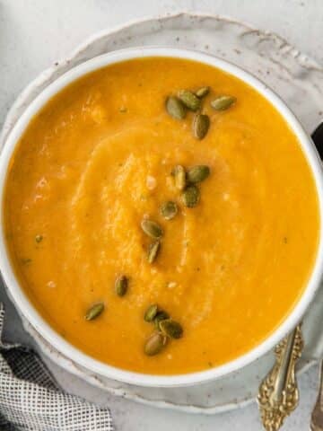 A bowl of butternut squash soup topped with pumpkin seeds, served on a plate with two vintage spoons and a checkered cloth nearby.