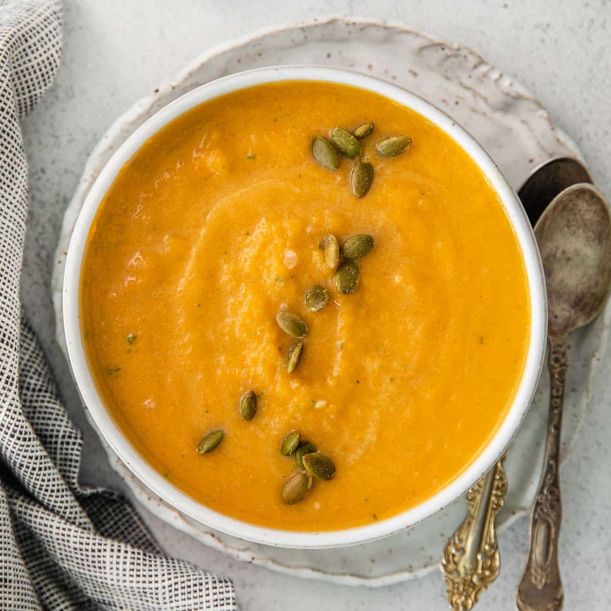 A bowl of butternut squash soup topped with pumpkin seeds, served on a plate with two vintage spoons and a checkered cloth nearby.