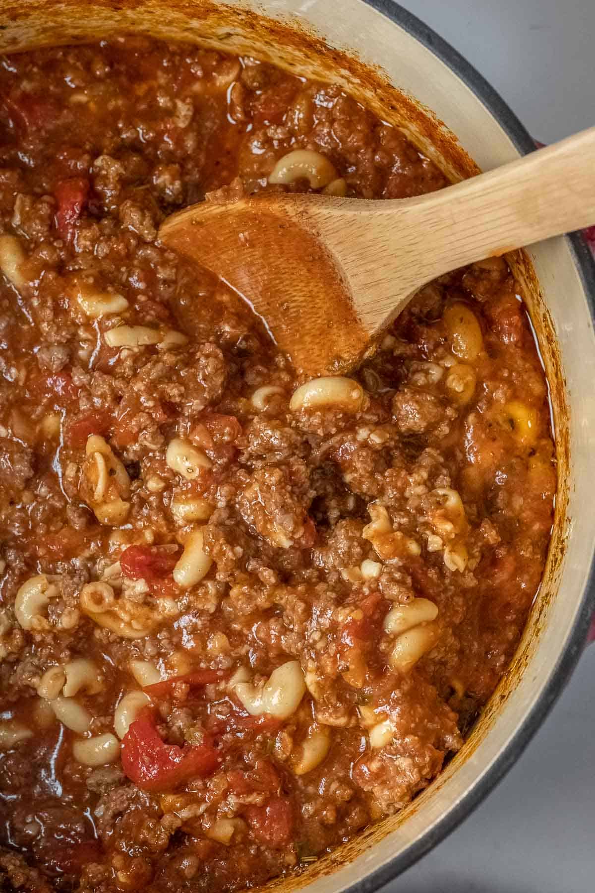 A pot of cooked goulash with ground beef, macaroni noodles, tomatoes, and sauce, being stirred with a wooden spoon.