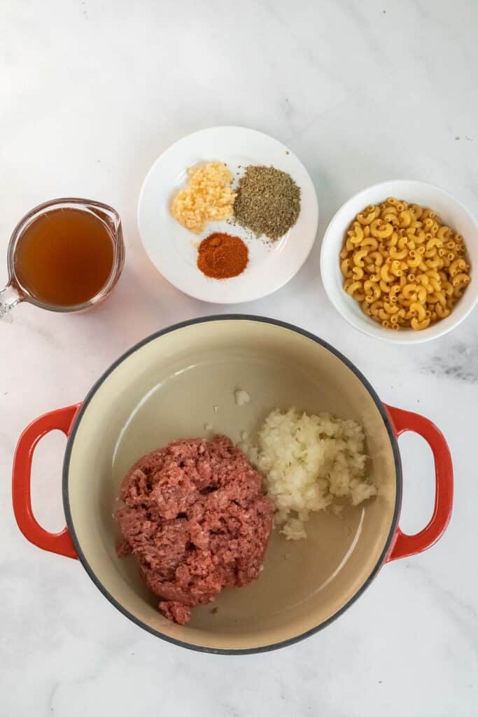 dutch oven with ground meat and chopped onion, next to bowls of elbow macaroni, spices, minced garlic, and a cup of broth on a white countertop.