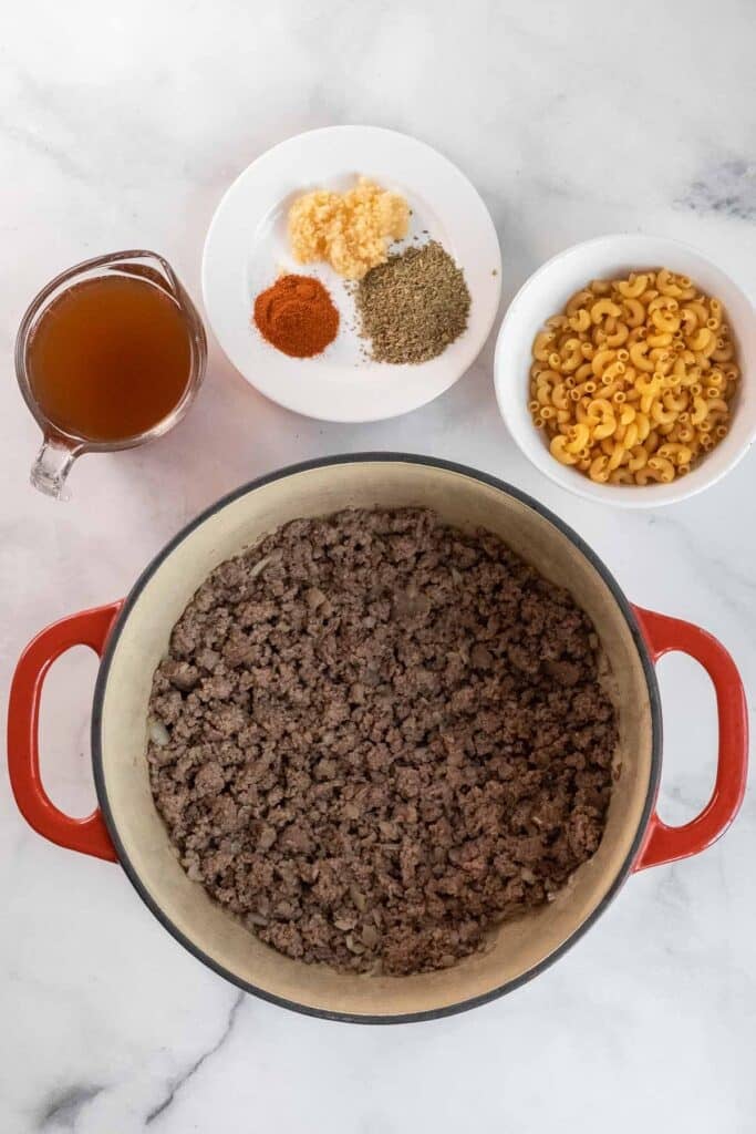 cooked ground beef in a dutch oven, uncooked macaroni in a bowl, broth in a measuring cup, and spices with minced garlic on a small white plate.