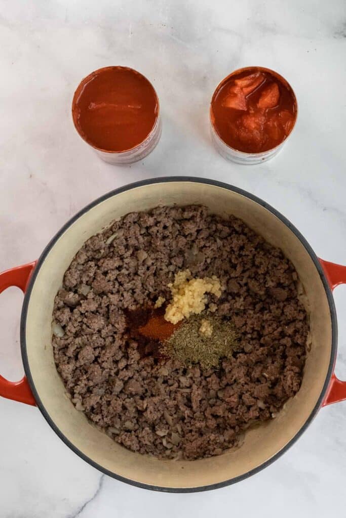 A dutch oven pot of cooked ground beef with spices and minced garlic, next to two open cans of tomato sauce and diced tomatoes on a marble surface.