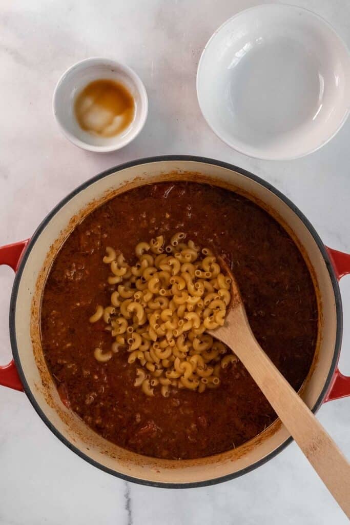 A red dutch oven filled with a meat sauce and uncooked elbow macaroni, being stirred with a wooden spoon; an empty bowl and a small dish are nearby.