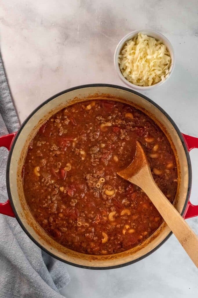 A large dutch oven of full off American goulash recipe with a wooden spoon, next to a small bowl of shredded cheese on a marble surface.