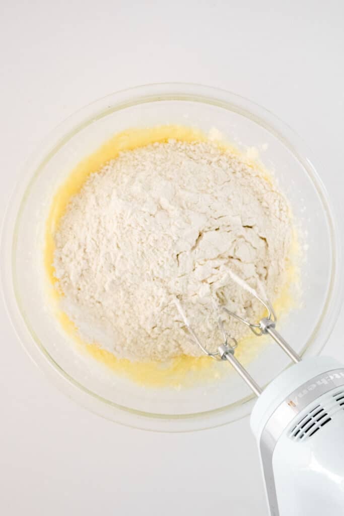 A mixing bowl with flour and yellow batter being mixed with an electric hand mixer on a white surface.