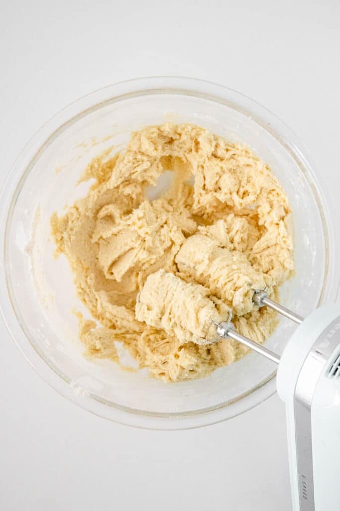 A glass bowl containing mixed cookie dough and a hand mixer with dough on the beaters, viewed from above.