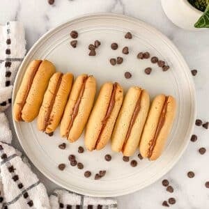 A plate with six milano cookies filled with chocolate ganache, surrounded by scattered chocolate chips on a marble surface.