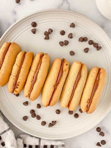 A plate with six milano cookies filled with chocolate ganache, surrounded by scattered chocolate chips on a marble surface.