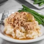 A plate with slow cooker mississippi chicken over mashed potatoes served with green beans. A slow cooker and a plate of green beans are in the background with a fork laying aside.