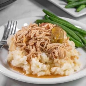 A plate with slow cooker mississippi chicken over mashed potatoes served with green beans. A slow cooker and a plate of green beans are in the background with a fork laying aside.