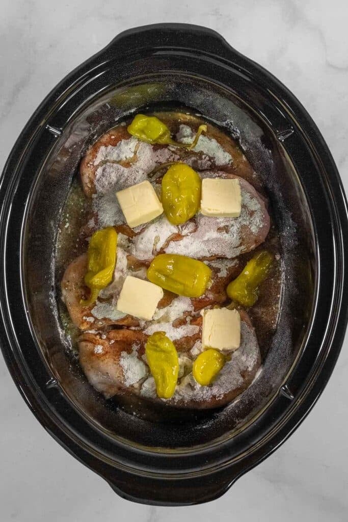 Raw seasoned chicken in a slow cooker, topped with butter slices and pepperoncini peppers, ready to be cooked.