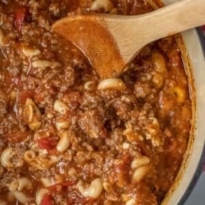 A close-up of an easy goulash recipe in a thick, tomatoes sauce being stirred with a wooden spoon in a pot.