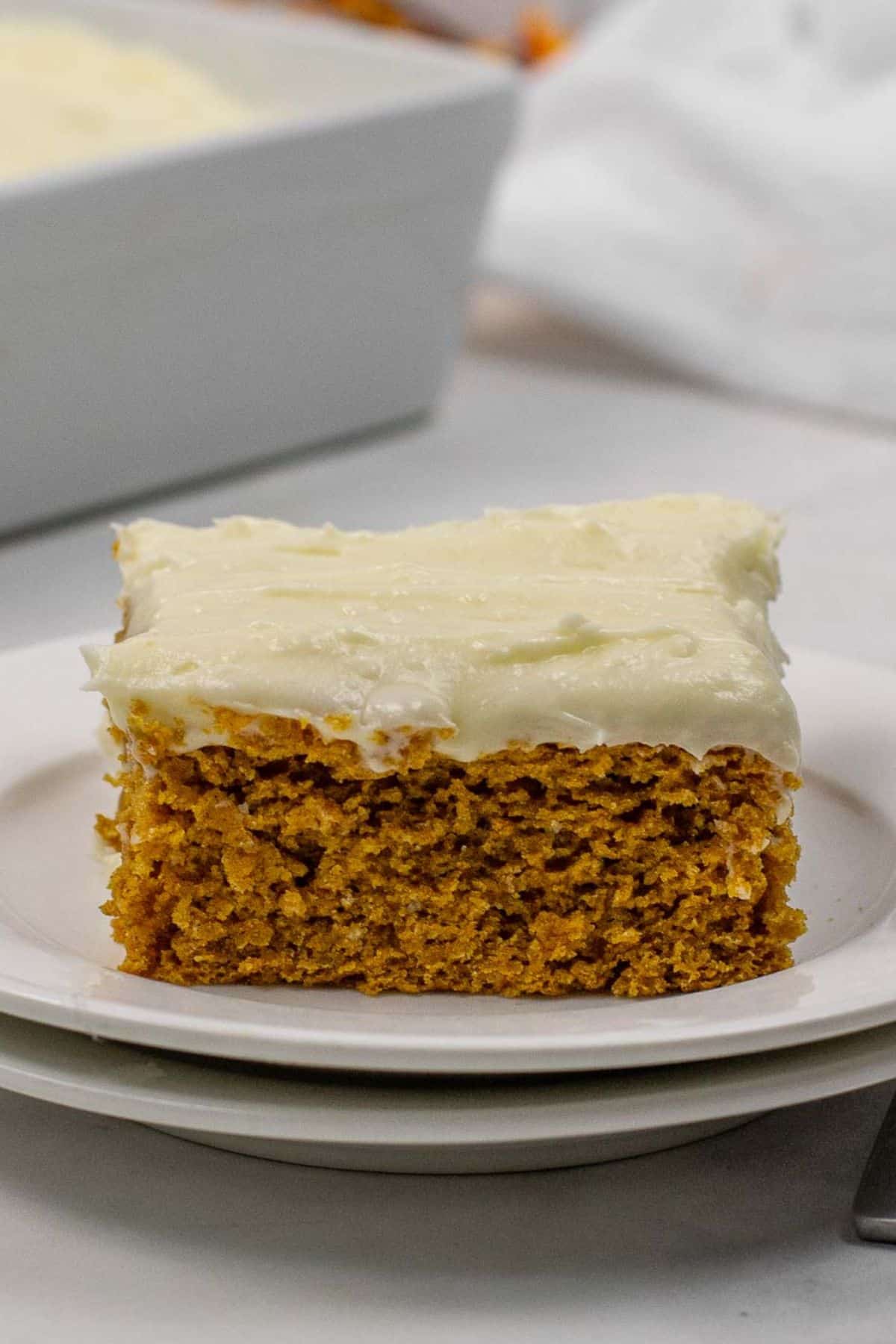 A square slice of pumpkin spice cake with cream cheese frosting sits on a white plate, with a baking dish in the background.