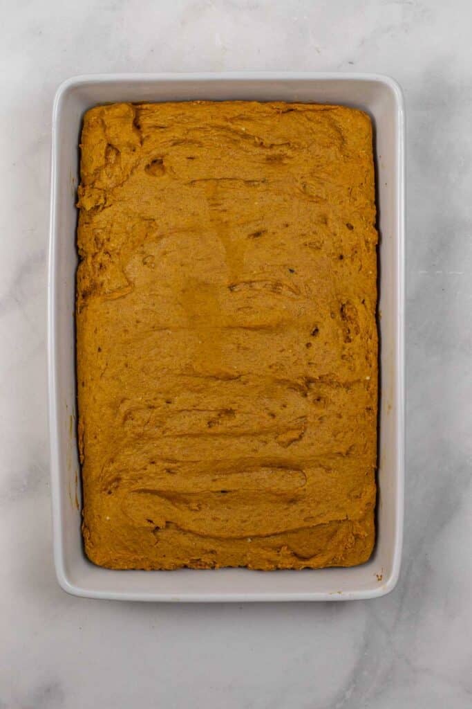 A rectangular baking dish filled with a baked, smooth, golden-brown pumpkin cake sits on a white marble surface.
