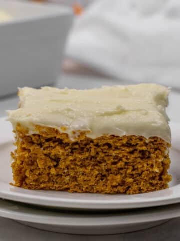 A slice of pumpkin cake with cream cheese frosting on a white plate, next to a fork.